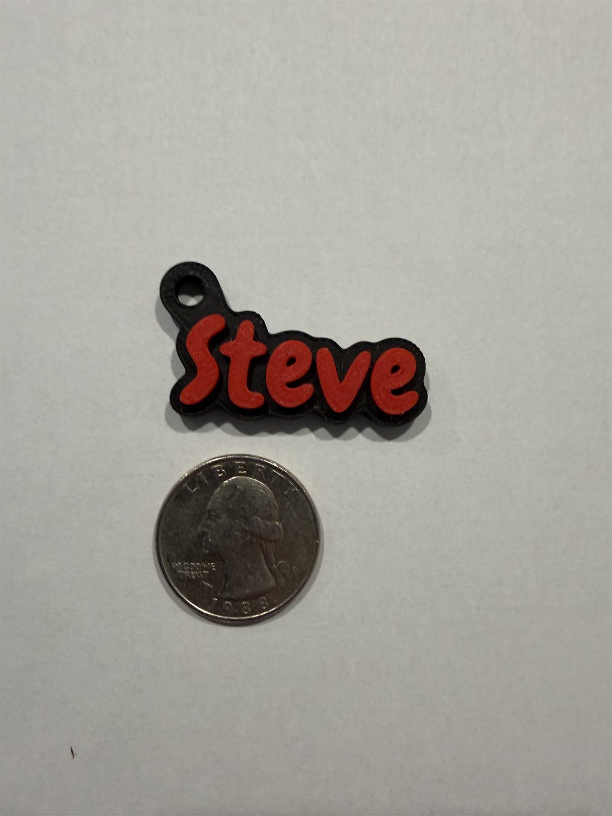 A red and black 'Steve' keychain next to a U.S. quarter for size reference