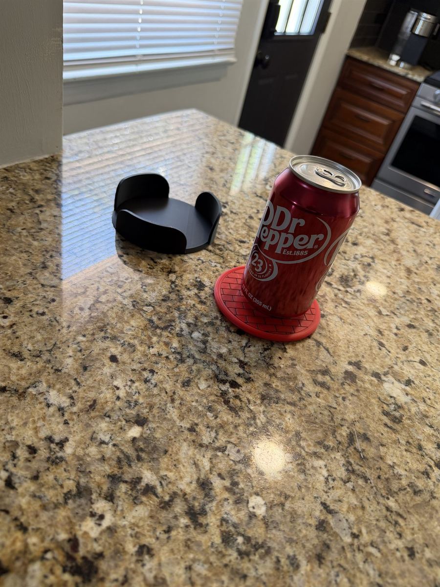 A red coaster holding a full soda can on a granite counter, with its holder nearby
